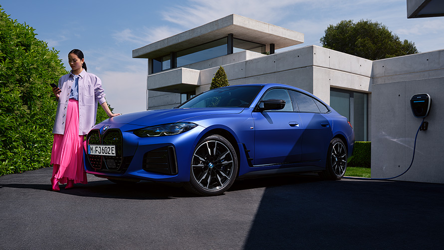 BMW i4 M50 G26 2021 BMW Individual Frozen Portimao Blau metallic, vue de trois quarts avant avec femme devant une maison